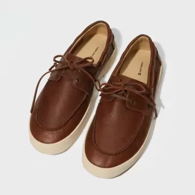 Sapatilhas castanhas com atacadores e sola branca sobre fundo branco.