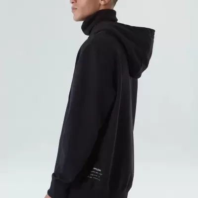 Homem com sweatshirt preta de capuz e gola alta com texto branco, calças pretas com riscas brancas, fundo cinza claro