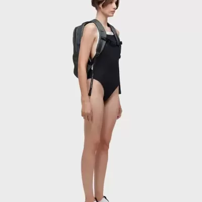 Modelo com fato de banho preto, mochila cinzenta e sapatos brancos e pretos em fundo branco