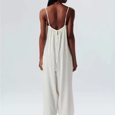 Macacão feminino branco com alças finas e cordão atrás, chapéu bege de palha, sandálias pretas.