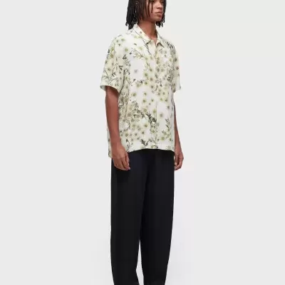 Homem a vestir camisa floral de manga curta, calças pretas largas e sandálias castanhas num fundo cinza claro