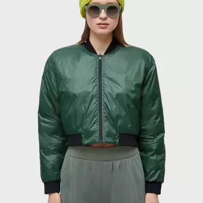 Mulher de casaco bomber verde escuro, calças cinza, gorro amarelo e óculos de sol