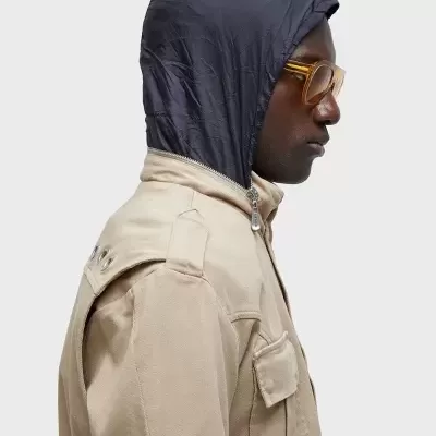 Homem com casaco bege e capuz preto, óculos amarelos, vista lateral