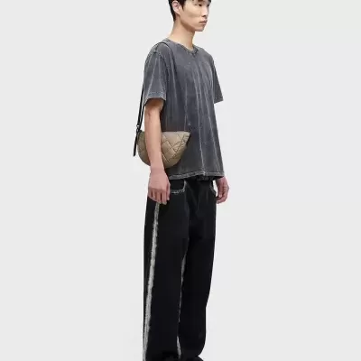 Homem com t-shirt cinza, calças pretas largas com faixa branca, ténis e bolsa bege matelassê.