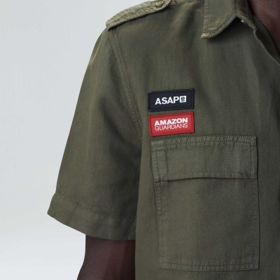 Camisa verde-oliva com dois emblemas e bolso frontal numa pessoa.