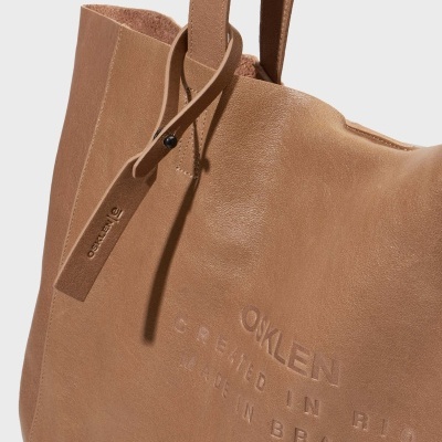 Mala tote castanha de couro com texto em relevo e tag pendurada da marca OSKLEN