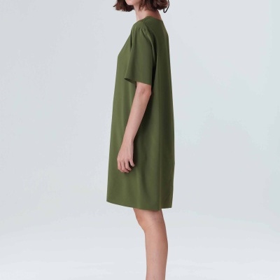 Mulher de perfil com vestido verde, chapéu verde e sandálias creme num fundo branco.