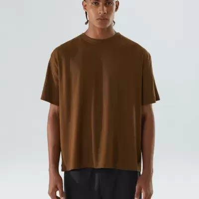 Homem a usar t-shirt castanha de manga curta com calças pretas, fundo cinza.