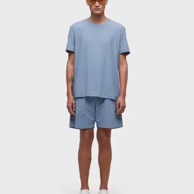 Homem modelo com conjunto azul de t-shirt e calções, sapatos brancos e óculos de sol laranja em fundo branco.