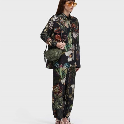 Mulher com camisa e calças pretas com padrão floral colorido, óculos de sol amarelos, sandálias brancas e mala verde texturizada em fundo branco