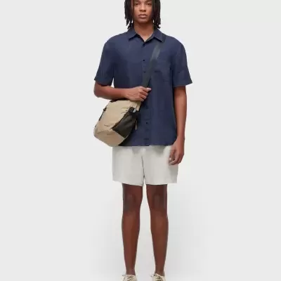 Pessoa com camisa azul escura, calções bege, ténis bege e mochila tiracolo OSKLEN