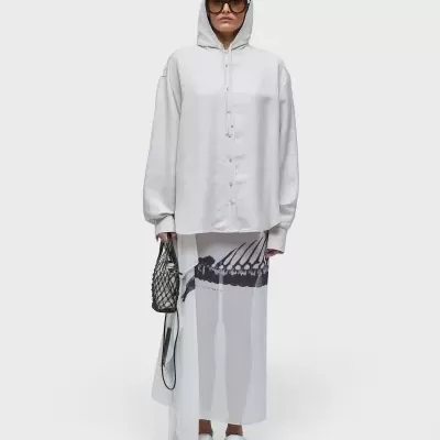 Mulher com hoodie branco, saia longa estampada, calça branca, mala preta e óculos escuros