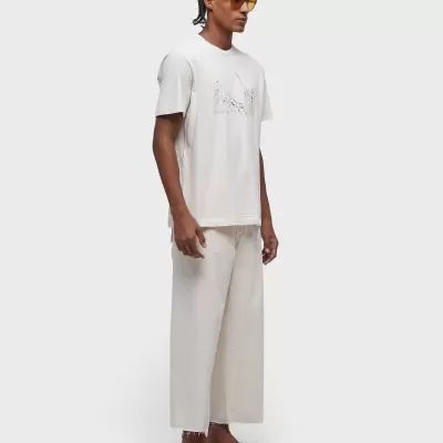 Homem com t-shirt branca e calças largas claras em fundo cinza.