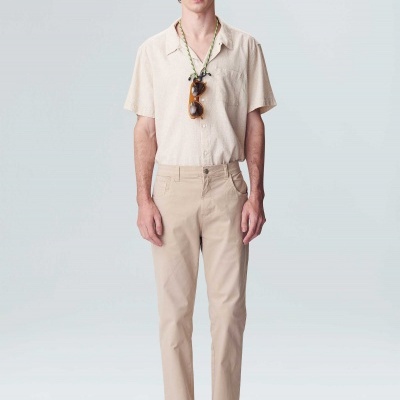 Homem de pé com camisa bege de manga curta, calças bege e sandálias pretas