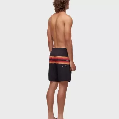 Homem com bermuda de banho preta com riscas coloridas e sandálias pretas num fundo branco.