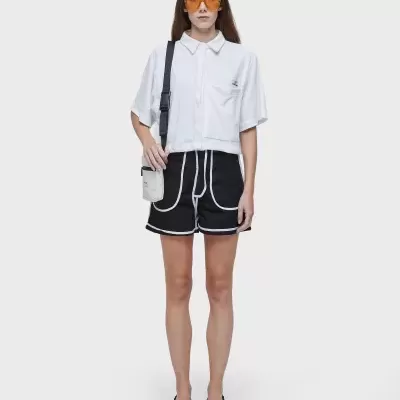 Mulher vestida com camisa branca, calções pretos com contorno branco, chinelos, bolsa branca e óculos de sol amarelos.