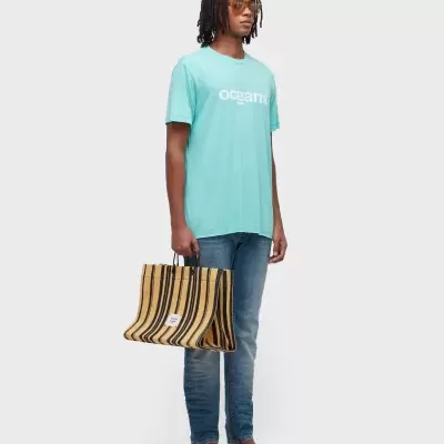 Homem com camiseta azul claro, jeans azuis, sandálias castanhas e mala listrada castanho e preto