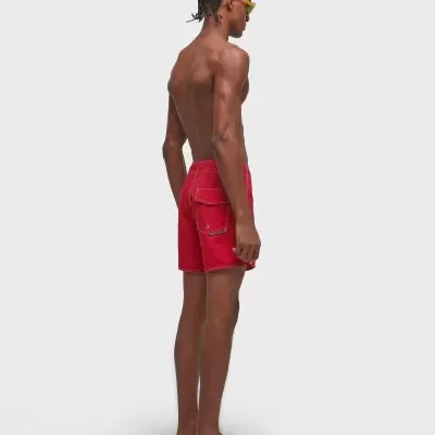 Homem a usar calções de banho vermelhos, óculos de sol amarelos e chinelos pretos num fundo branco.