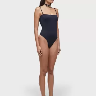 Fato de banho feminino preto com alças finas usado por mulher com acessórios pretos e fundo cinza