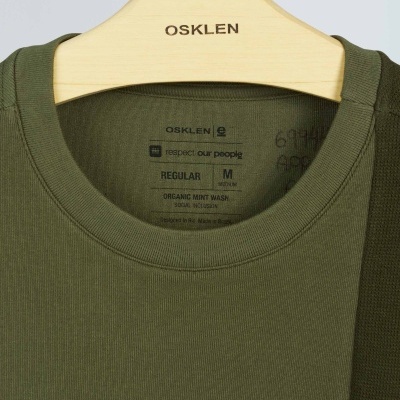 T-shirt verde-oliva com etiqueta interna OSKLEN pendurada em cabide de madeira