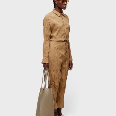 Pessoa com macacão castanho e óculos de sol a segurar mala bege.