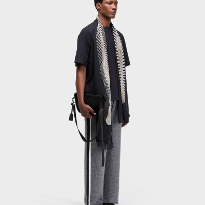 Homem com camisola preta, cachecol preto com padrão branco, calças cinzentas com risca branca lateral e carteira preta na mão direita