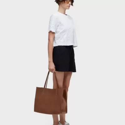 Mulher de perfil com t-shirt branca, calções pretos, sapatilhas brancas e mala castanha de couro