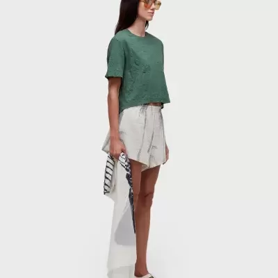 Mulher vestindo roupas casuais com camisola verde, calções e calçado branco, segurando lenço branco com padrão.