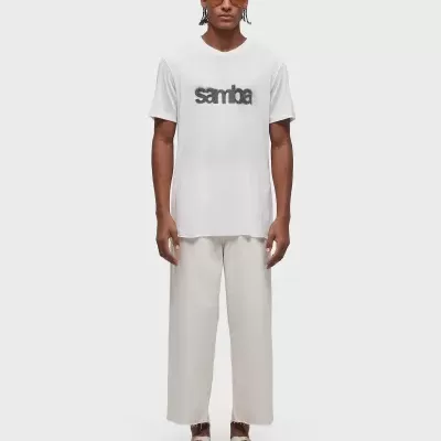 Homem com t-shirt branca com a palavra 'samba', calças largas brancas e sapatilhas claras.