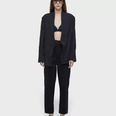 Mulher com fato preto, top preto, sandálias pretas e óculos de sol castanhos sobre fundo cinza