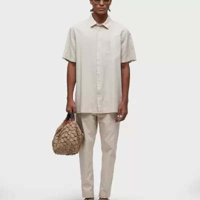 Homem com camisa e calças bege, óculos de sol amarelos, mala texturizada castanha e sapatos castanhos