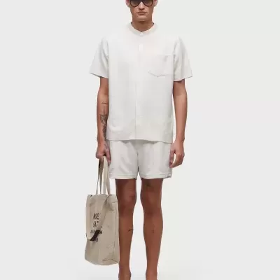 Homem com conjunto branco de camisa e calções, óculos escuros, sandálias pretas e mala bege com texto 'MC'.