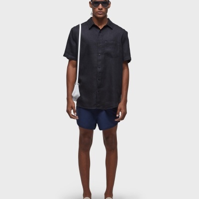 Homem com camisa preta, calções azuis, ténis bege e óculos escuros, bolsa branca sobre o ombro