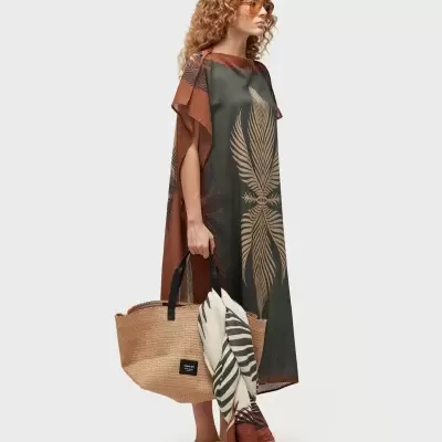 Mulher com vestido comprido com padrão de folhas, mala de palha com lenço e sapatos bege