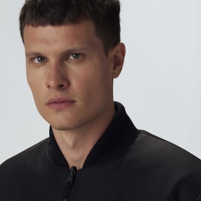 Homem com jaqueta preta de colarinho tipo bomber em fundo cinza claro.