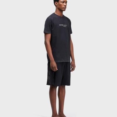 Homem de pé com t-shirt preta com texto branco, calções pretos com padrão lateral e chinelos pretos