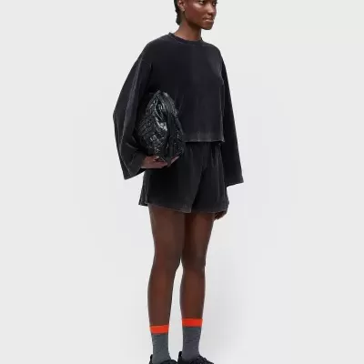 Mulher vestindo casaco e calções pretos, meias cinza com risca laranja, ténis pretos e segurando mala preta