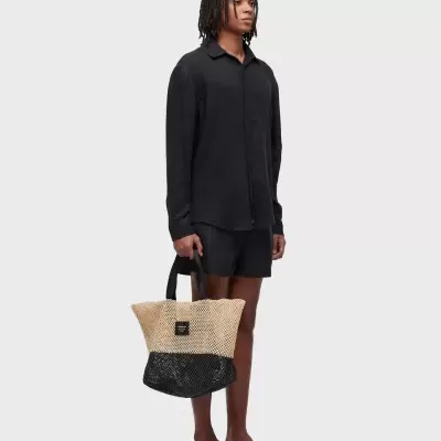 Homem com camisa preta, calções pretos, chinelos e mala tote tricolor bege e preta