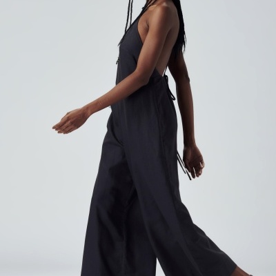 Macacão preto de tecido leve com alças finas e calças largas usado por mulher com sandálias pretas e óculos escuros