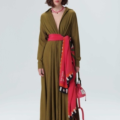 Mulher com vestido longo verde-oliva, cinto e echarpe vermelhos, mala pequena e sandálias pretas.