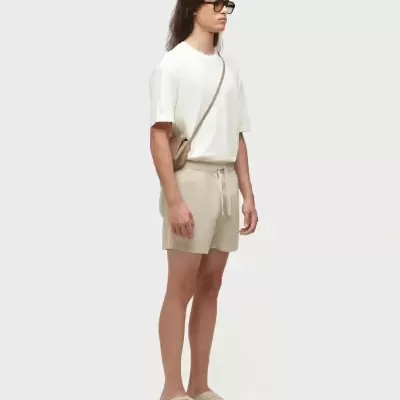 Homem jovem com t-shirt branca, calções bege, sapatos slip-on trançados, óculos escuros e mala bege a tiracolo