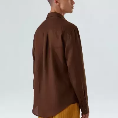 Pessoa de costas com camisa castanha de manga comprida e calças mostarda caneladas em fundo branco