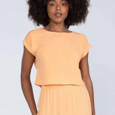 Mulher com conjunto cor de pêssego claro, t-shirt de manga curta e saia com pregas em fundo cinza claro