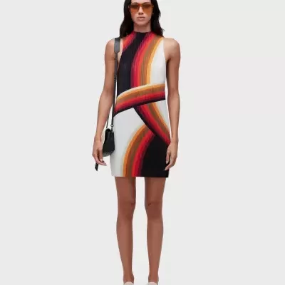 Mulher a usar vestido curto com padrão abstrato colorido, óculos de sol castanhos, bolsa preta e ténis brancos.