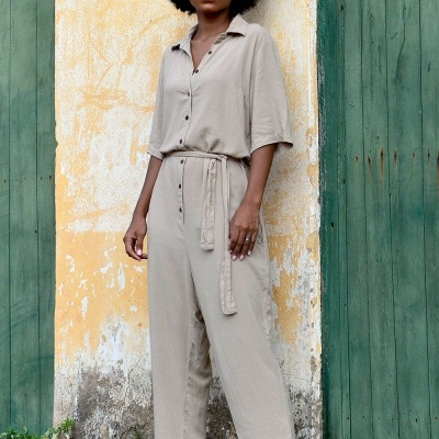 Mulher de macacão bege com cinto e sandálias verdes junto a uma parede amarela e portas verdes