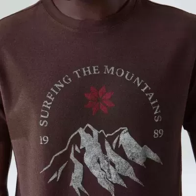 T-shirt castanha com estampado de montanhas, flor e texto em branco da marca Osklen.