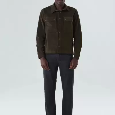 Homem com camisa castanha de camurça, calças pretas e sapatos pretos em fundo branco