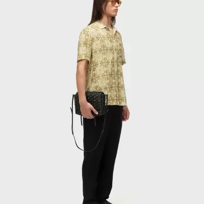 Homem com camisa estampada amarela, calças pretas, sandálias e bolsa pretas em fundo branco.