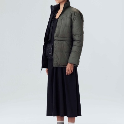 Mulher com casaco acolchoado verde-oliva, vestido preto e botas brancas numa pose lateral