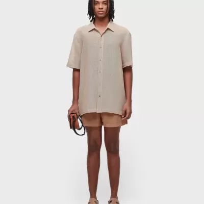 Pessoa com camisa bege de manga curta, calções castanhos, sandálias e bolsa castanha